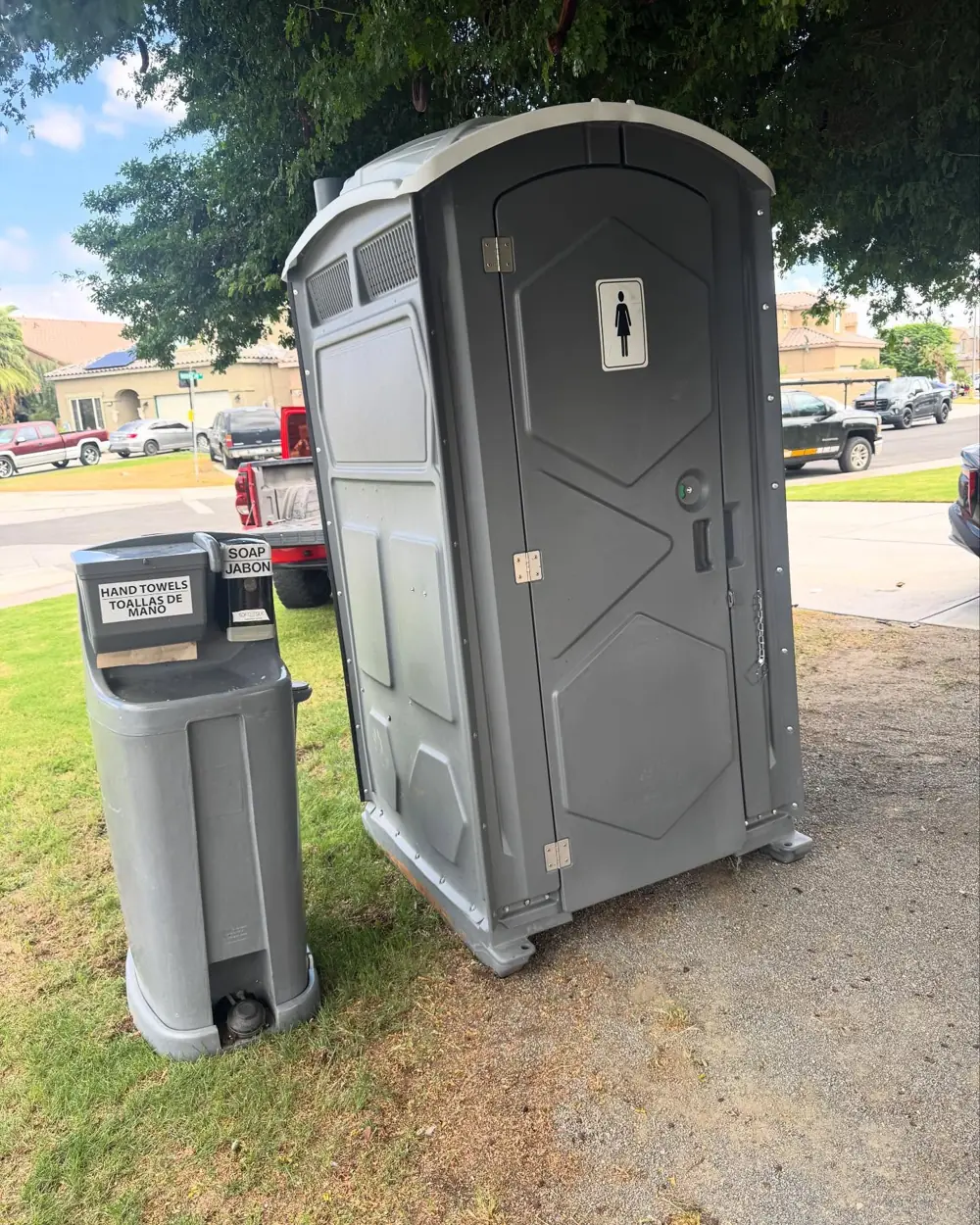 Gray portable toilet beside a two-sink foot-pump hand-washing station on grass